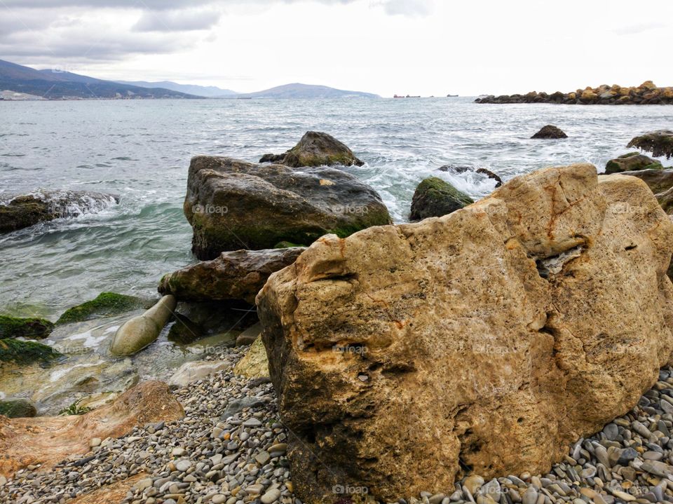 Stone beach, boulders. Rainy weather. Waves break on rocks and rocks. In the background is a mountain range. The barges are floating.