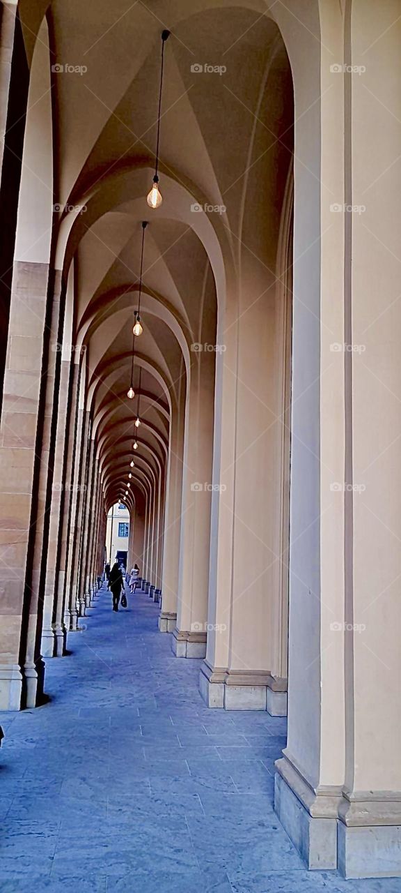 High arches are covering public walkways in the city of „Munich“, „Bavaria“ as one of many signs of an antique traditional influence on the architecture everywhere. Small street lamps hang high above the pedestrians. 2022. Hypnotic Productions