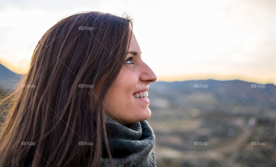 Profile portrait of young caucasian girl smiling against sunset over mountains.