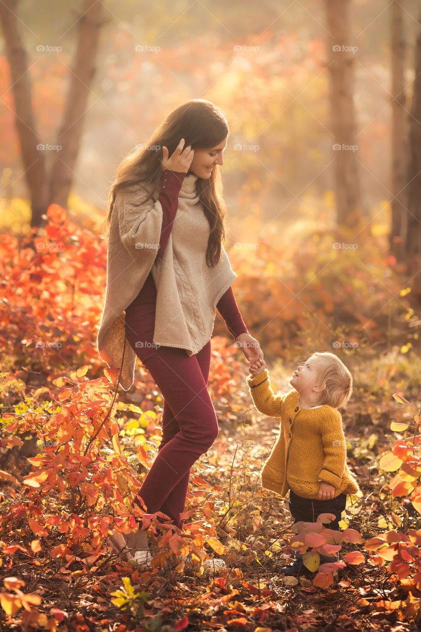 Young mother with cute baby daughter in knitted wear walking in warm and cozy autumn forest 