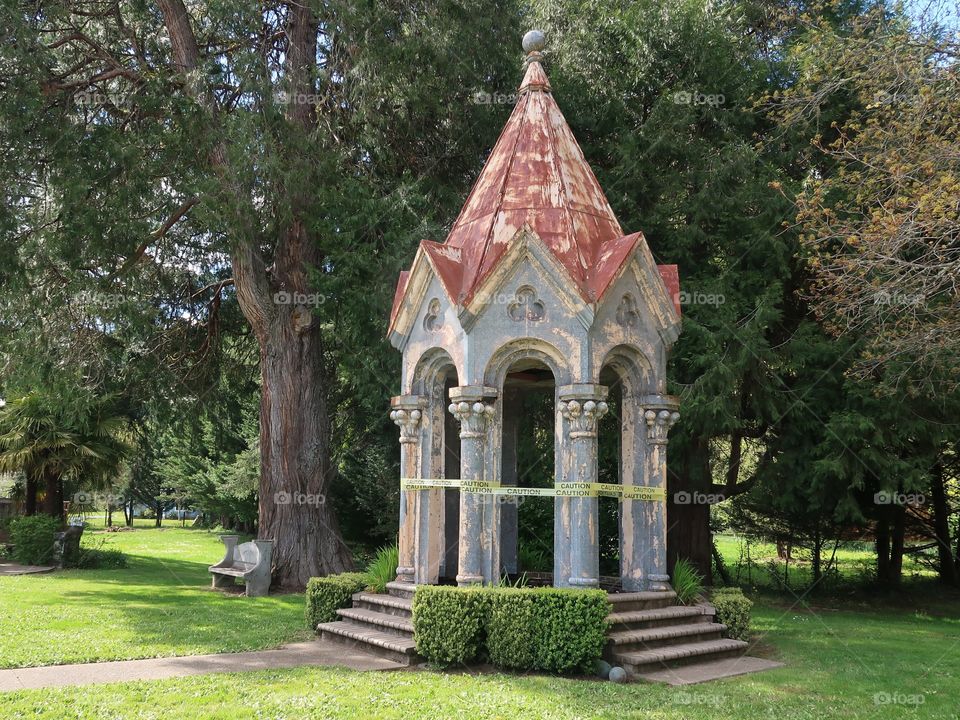 A large decorative stone gazebo in a nice park that has developed a lean and is in need of repair. 