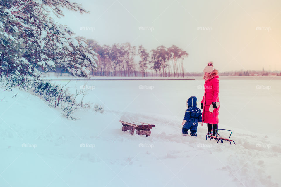 Mother and her little daughter are spending time together walking outdoors in forest in winter while snow falling. Woman is pulling sled, a few years old girl is walking through the deep snow, enjoying wintertime