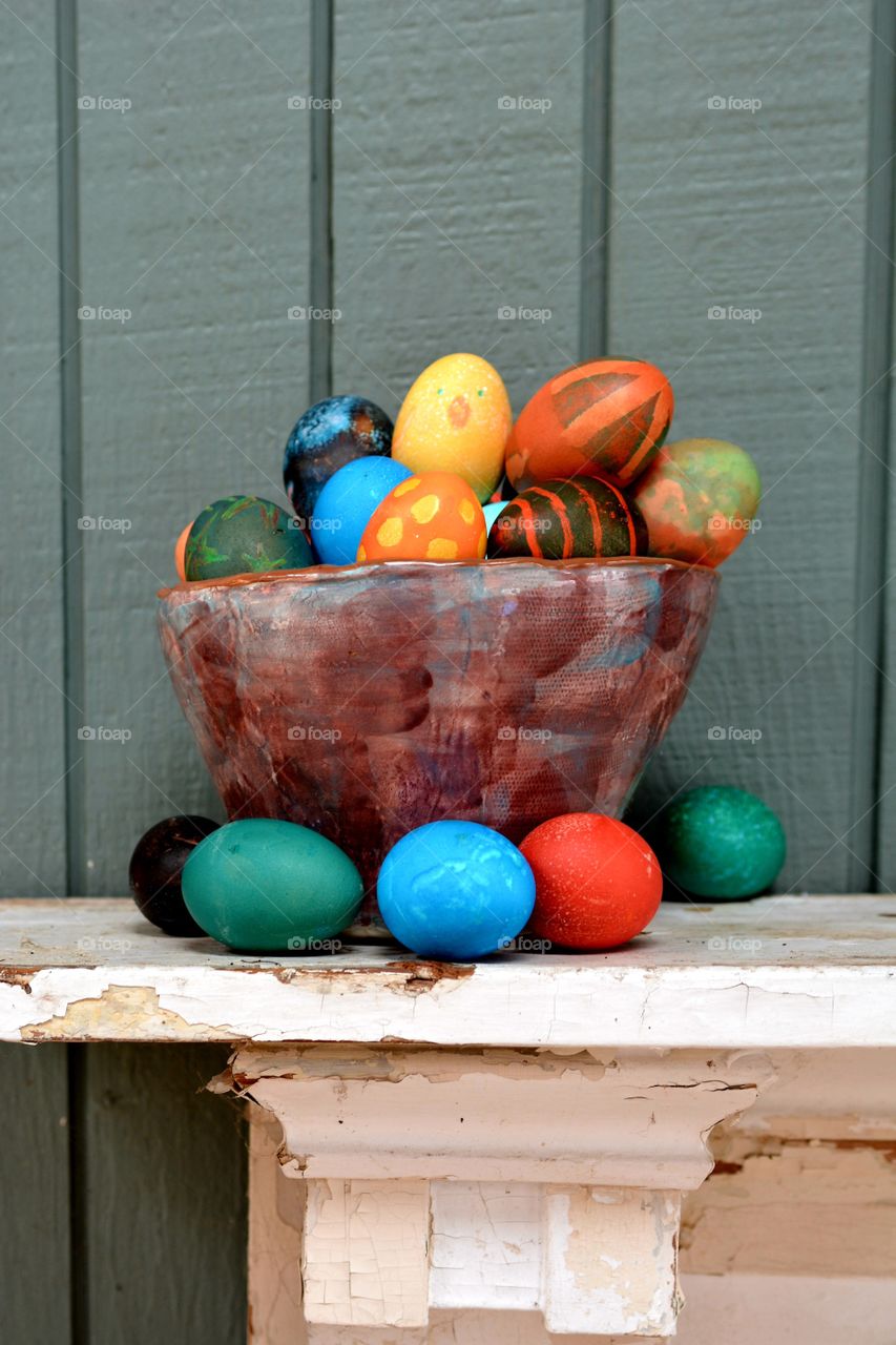 Deep colored vibrant Easter eggs are seen in a handmade ceramic bowl on a wooden, weathered white shelf