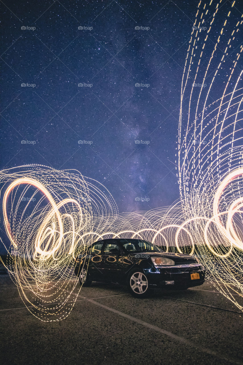 A black car circled by light painting on a dark summer night, with the Milky Way galactic core visible high up in the sky. 