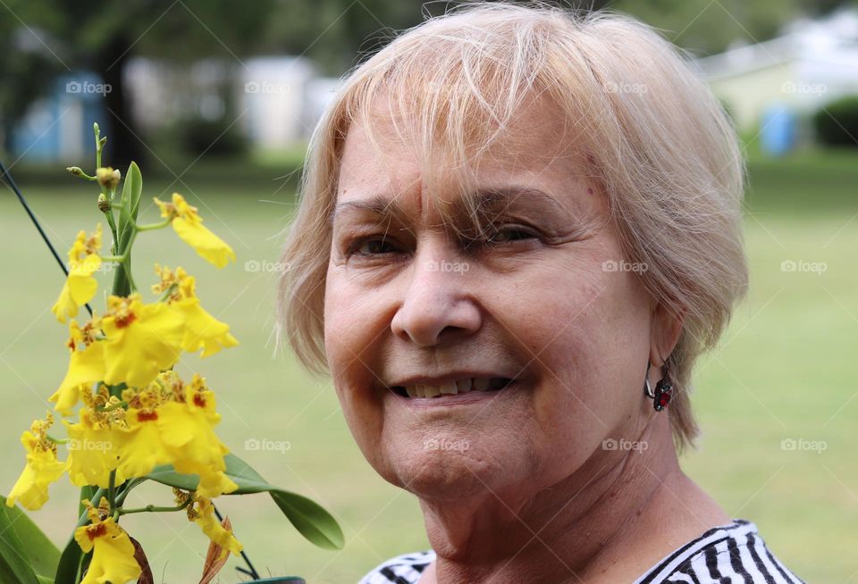 Closeup face happy expression senior woman outdoors next to flowering yellow orchid