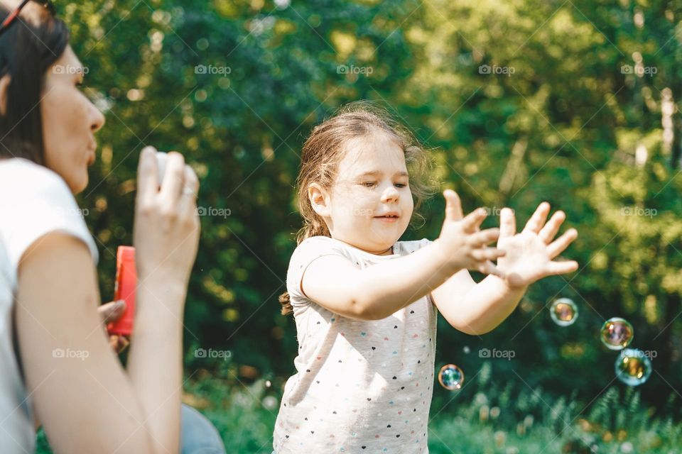 Cute toddler girl blows on soap bubbles with his mother in sunny summer day