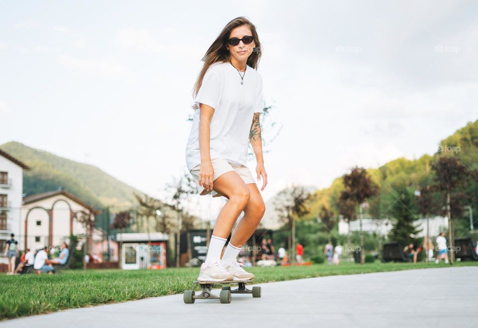 Slim young woman with long blonde hair in light sports clothes on longboard in outdoor skatepark at sunset time