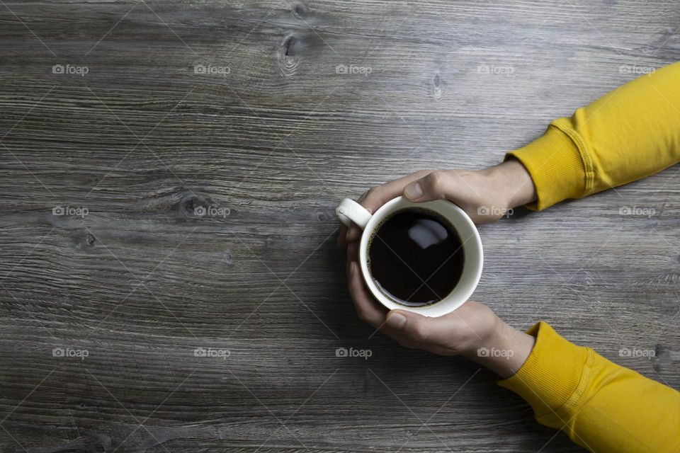 A man drinks coffee in the morning. Men's hands holding a white cup of coffee in a yellow jacket lie on a gray wooden surface, which is used as a background or a surface with incident light.