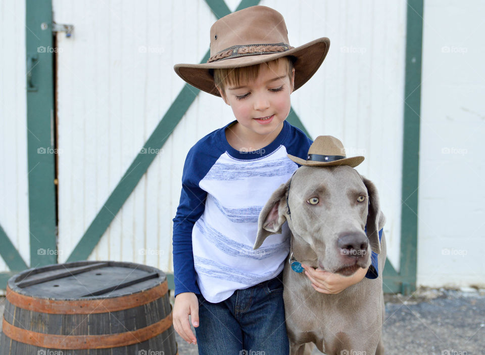 Young boy hugging his Weimaraner dog in front of a barn door outdoors and both wearing cowboy hats