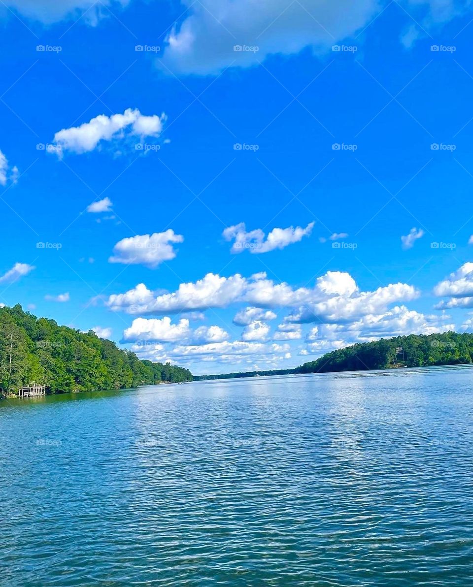 Puffy, white clouds float in a vibrant blue sky above the water of a like, lined by lush trees.