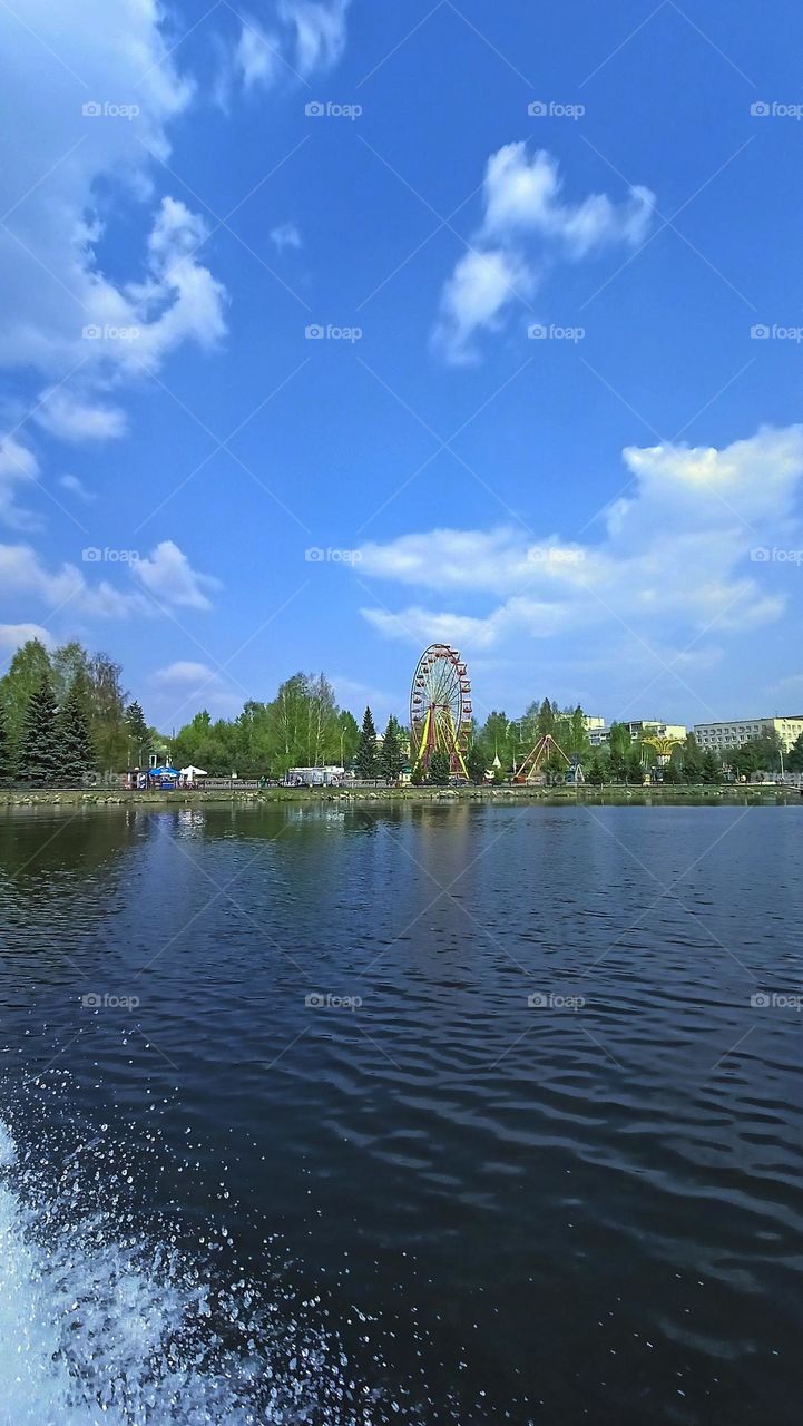 urban landscape, water, pond, view from the pond to the park, amusement park, attractions, ferris wheel, city, spring, blue sky, sunny day