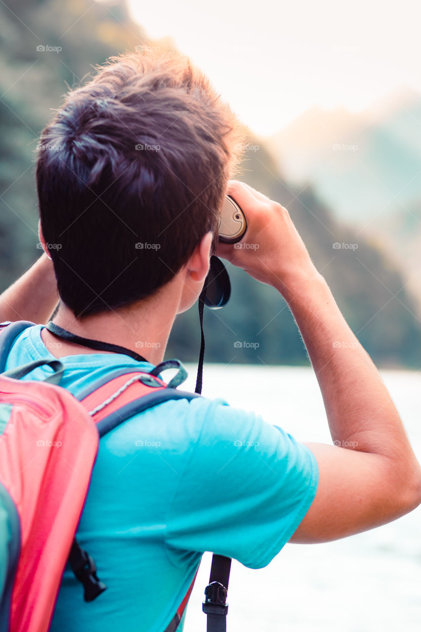 Young tourist with backpack looks through a binoculars on mountains peaks, stands on a rock over a river. Boy spends a vacation in mountains, wandering with backpack, he is wearing sports summer clothes