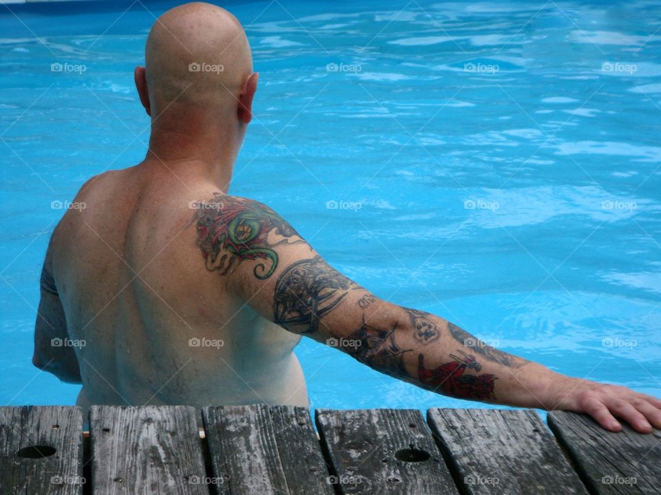 Summer activities in the USA. Man standing in waist deep backyard swimming pool. He's got an arm tattoo and is enjoying the sun!