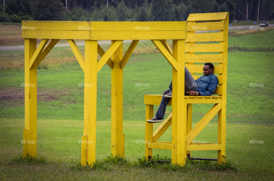 A man sits by an oversized yellow chair and table by a country road