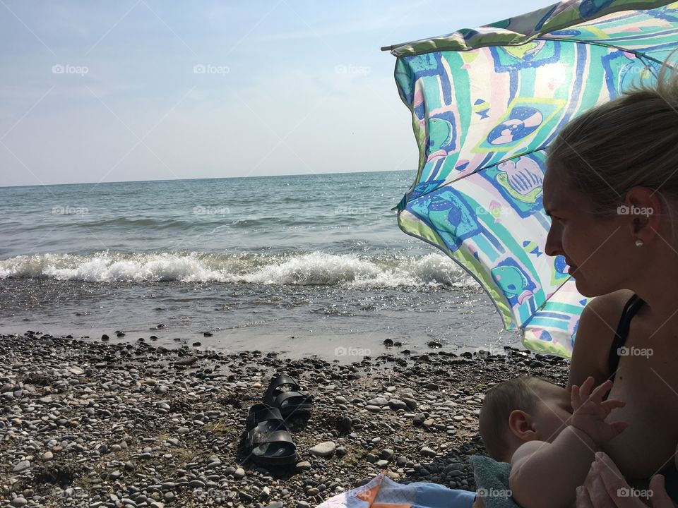 A young mother is nursing her infant peacefully on the beach, under protection of an umbrella, waves crashing near-by as she looks on into the distance under the sun and blue sky. Blue, green and neutral tones. 