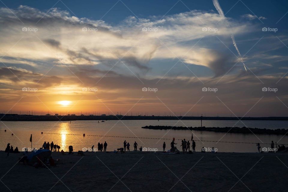 sunset on the sea in summer, expressive white clouds, blue sky, in the foreground the beach and silhouettes of people resting