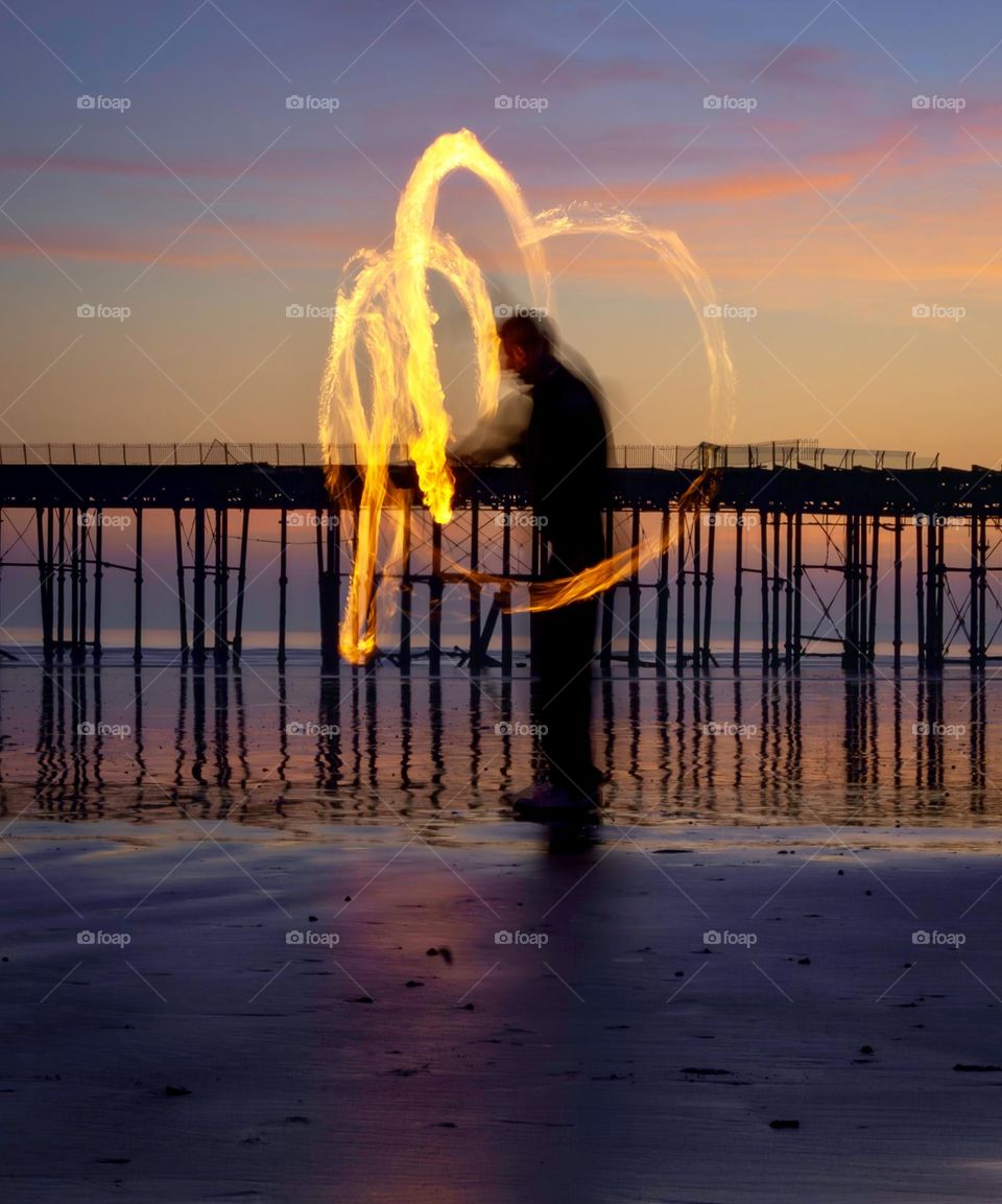 Man spinning fire poi on the beach, with an old pier in the background, around sunset