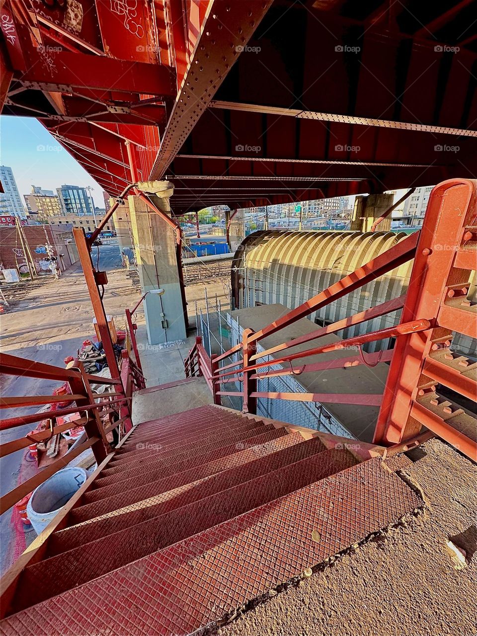 This is the red metal staircase of the „Pulaski Bridge“ at „Newtown Creek“ in LIC, Queens going down to street level. The warm light of the setting sun casts shadows onto its structures that section it off in unusual ways. 2024. Hypnotic Productions