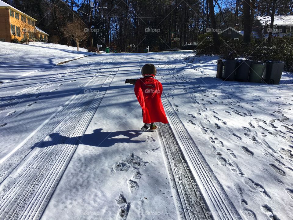 Little boy running in Superman red cape through snow covered streets.