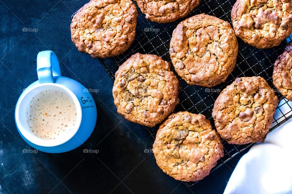 Chocolate chip cookies on a wire rack with a hot cappuccino in a blue mug at the side.