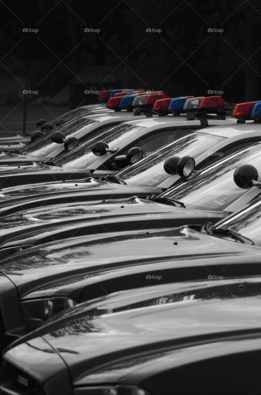 A fleet of black and white police cars with the red, white, and, blue berry lights are lined up on a residential street.