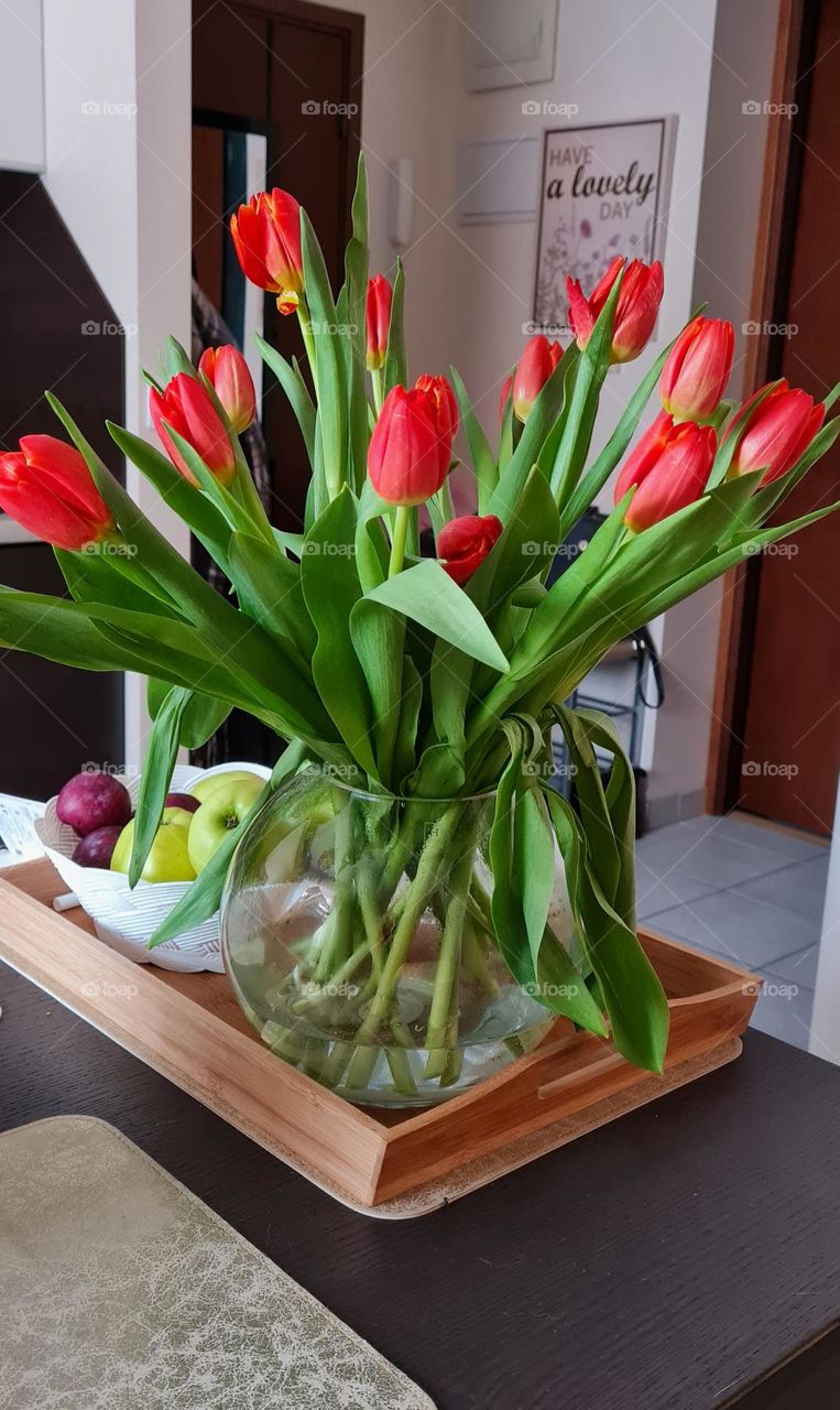 Many red tulips standing in a round transparent vase on the kitchen table. Lifestyle 