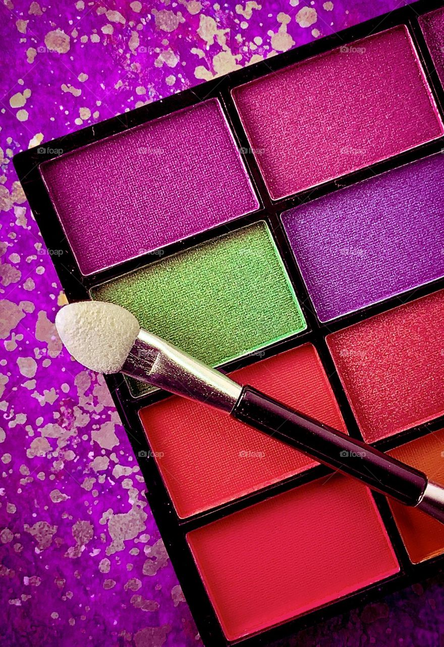 Make up palette in vibrant shades of reds and pink with one section in green, against a pink patterned background 