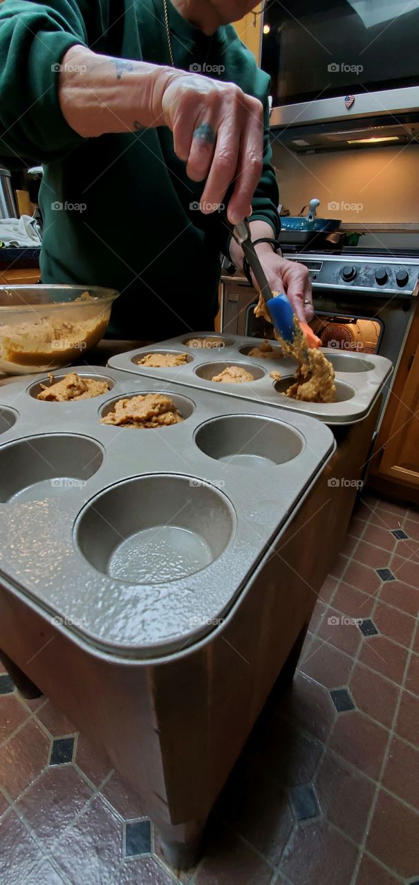 Homemade sweet potato muffins being spooned into muffin pans from glass bowl. Spatulas used to fill pan.