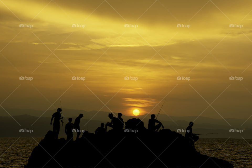 Silhouette of tourists Fishing on the rocks in sea and Golden light of sunrise behind the mountains.