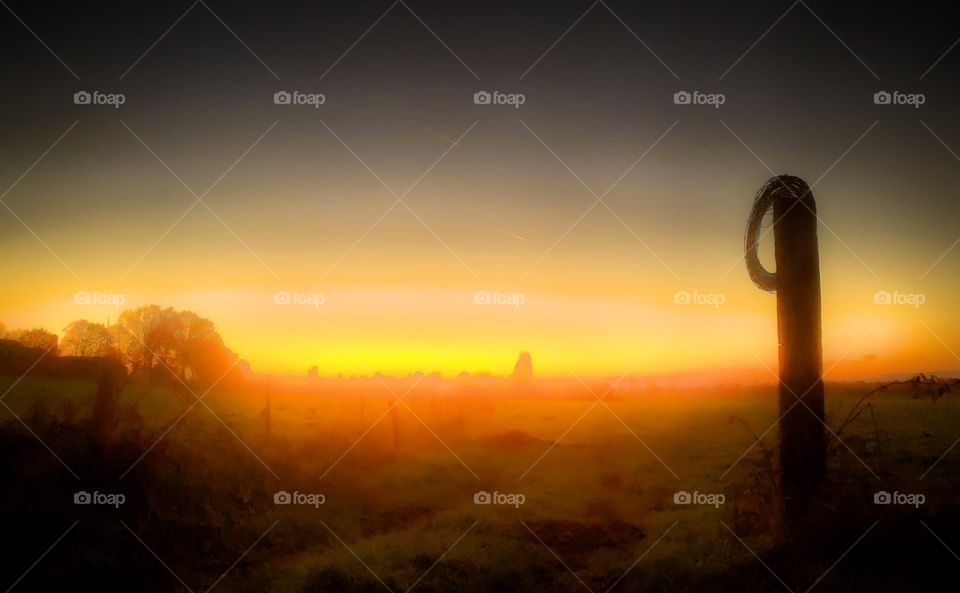 Fiery red orange glowing sunrise or sunset over a Grassy farmfield behind a barb wired post