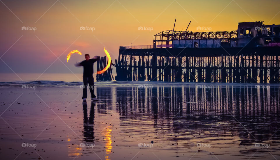 A man in warm clothing spins fire poi on the beach, just after sunset, against the backdrop of the burnt out remains of Hastings pier