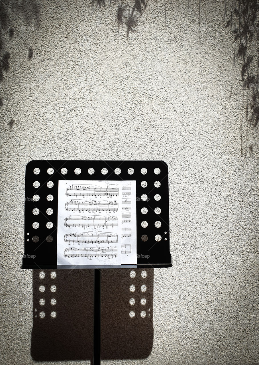 Black music stand with notes, casting an expressive shadow on the textured plastered wall on a bright sunny summer day