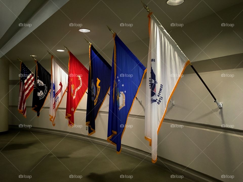 Collection of United States National, Prisoner of War and Missing in Action, Army, Marine Corps, Navy, Air Force, and Coast Guard Flags mounted on a white wall 