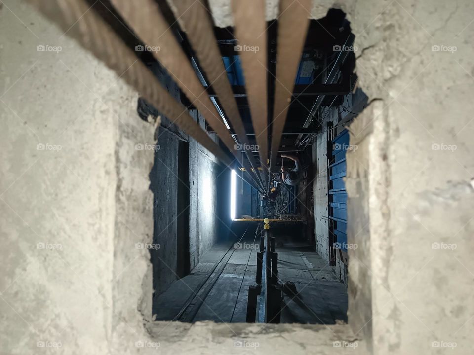 "Peering into the heart of the elevator shaft, where steel cables stretch endlessly upwards, tracing a path through the shadows. A worker, dwarfed by the towering walls, brings a human touch to the industrial depths."
