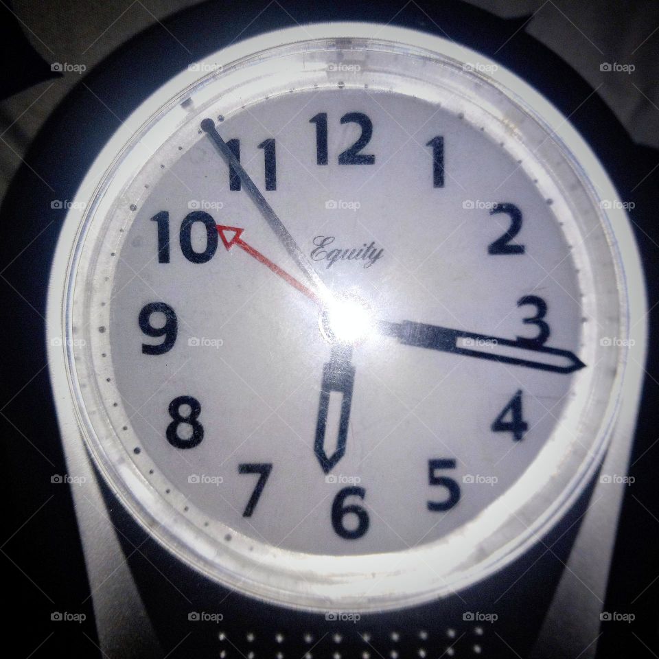 A bright white, round face of a clock with large black numbers, a red alarm arrow, black second hand and white outlined with black hands to reflect the time.