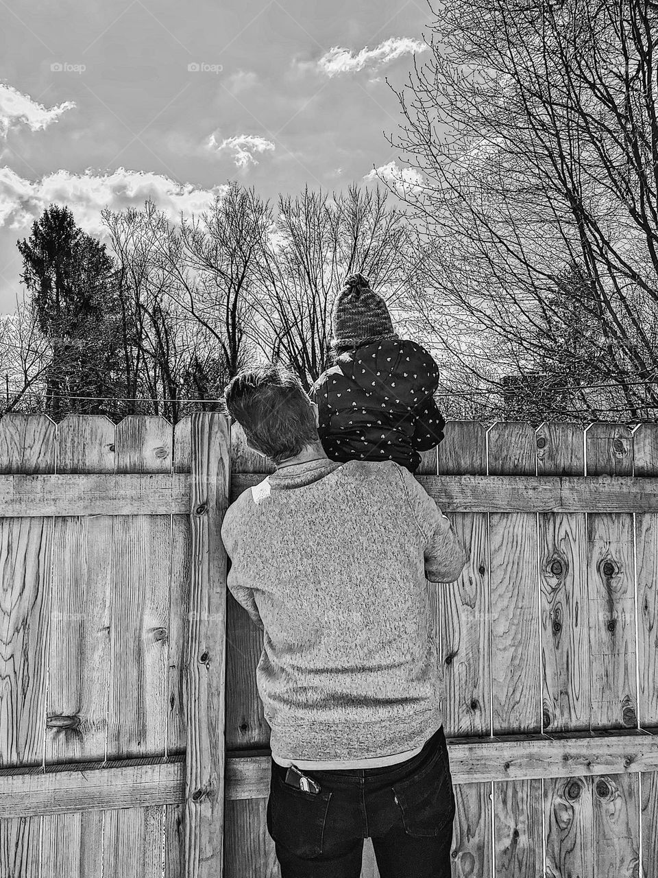 Toddler peering over fence while adult holds them up, Nosey neighbor toddler girl, curious toddler girl wants a better view of the neighbors, neighborhood watch toddler girl, toddler gets a better view atop the shoulder of an adult, monochrome pic 