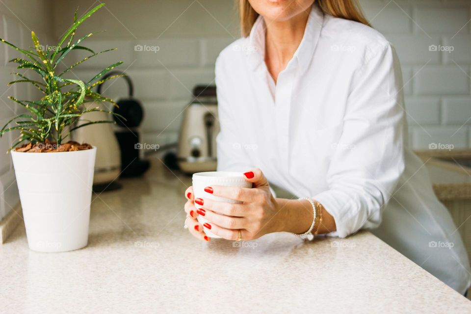 Crop photo of charming blonde woman with long fair hair in white shirt with morning cup of the tea in hands on bright kitchen before working day