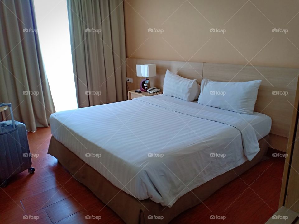 a comfortable king sized bed room in hotel with white color pillow, blanket, sheet. room facilities are side table, lampu, and phone. room Vincent design is wooden floor and wallpapers with blind curtain.
