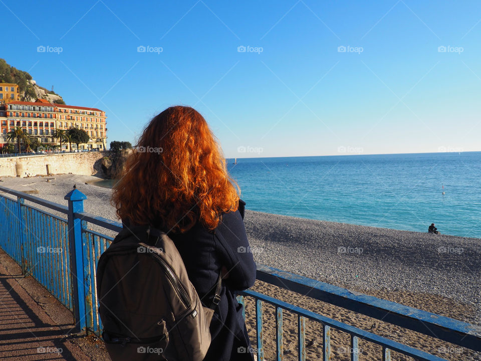 Redhead young woman looking out onto the beach from the Promenade des Anglais in Nice, France.