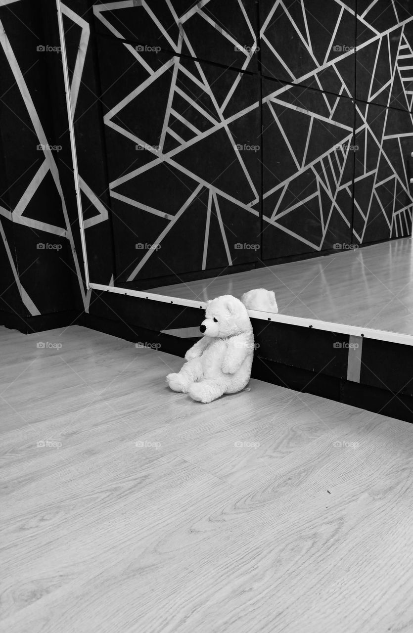 a white bear sits alone on the floor by the mirror. white and fluffy soft toy. Black and white. black and white photo. lines. mirror.