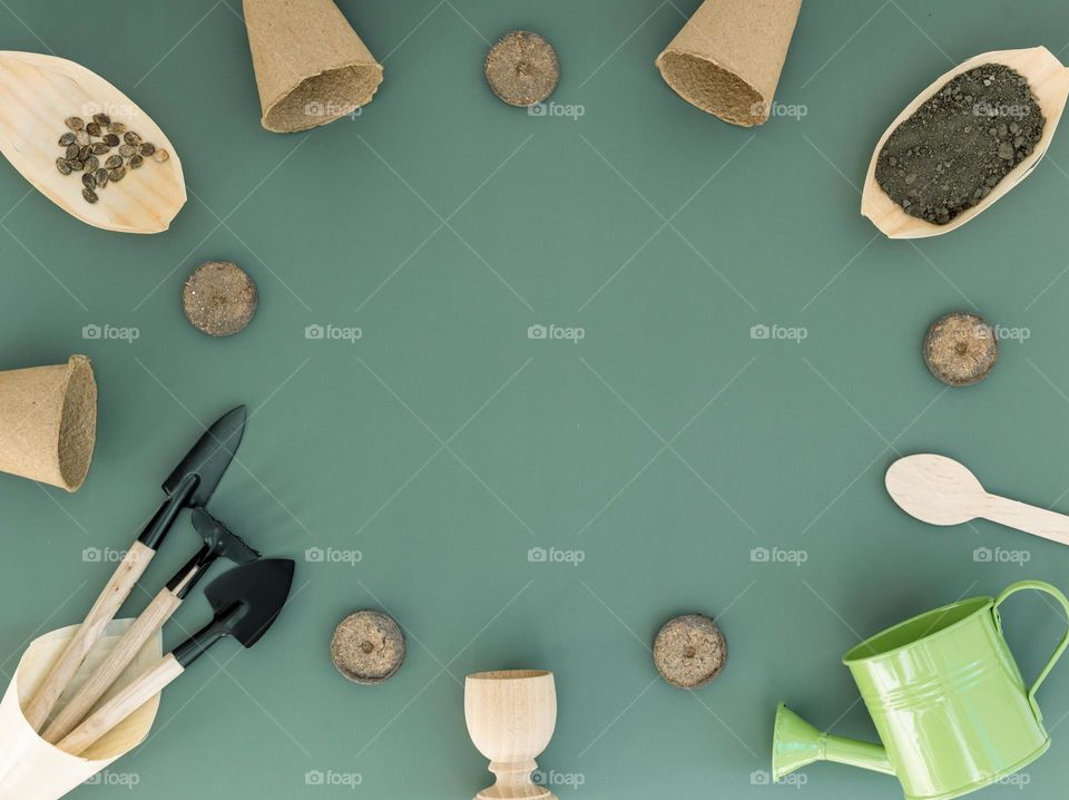 Three gardening tools, cardboard mills, wooden boats with soil, waterbase seeds and a green watering can are arranged in an oval frame on a natural green light with copy space in the center, flat lay close-up.