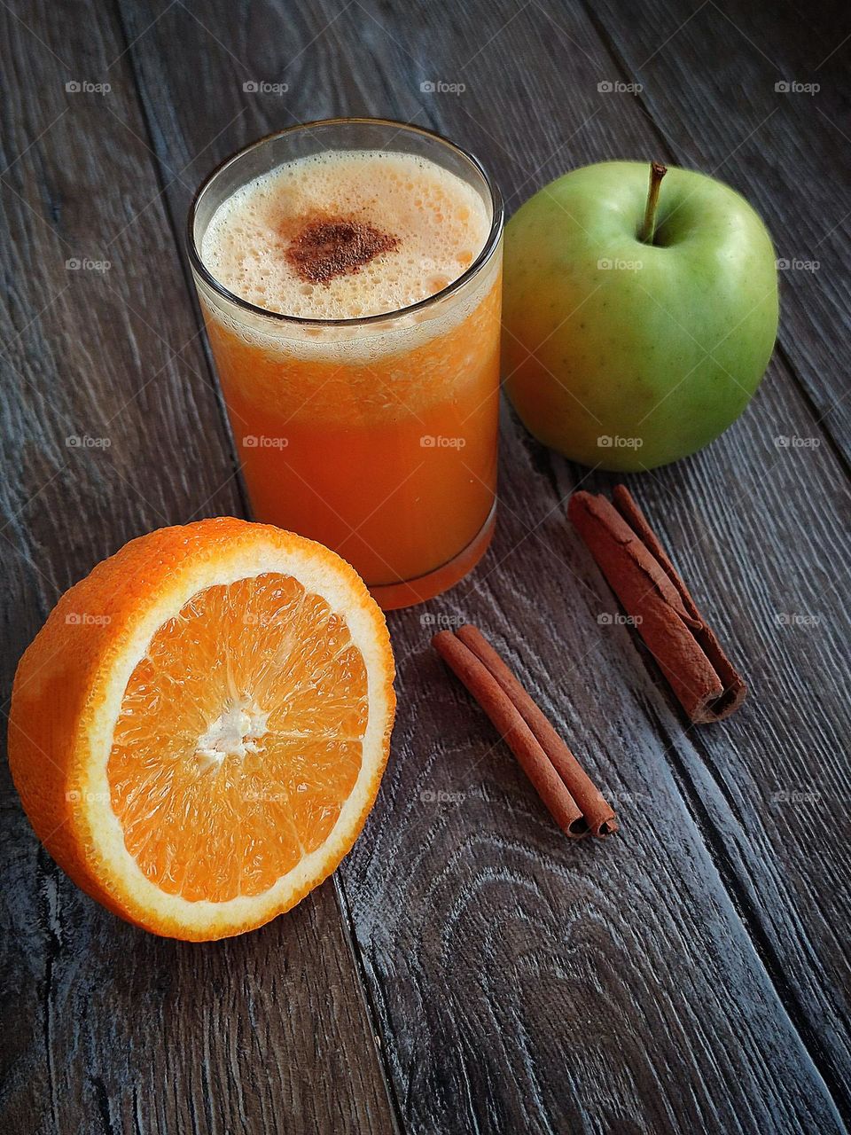On a wooden surface stands a glass with an orange smoothie, the surface of which is decorated with ground cinnamon in the form of a heart. Ingredients lie nearby: half an orange, a green apple and two cinnamon sticks