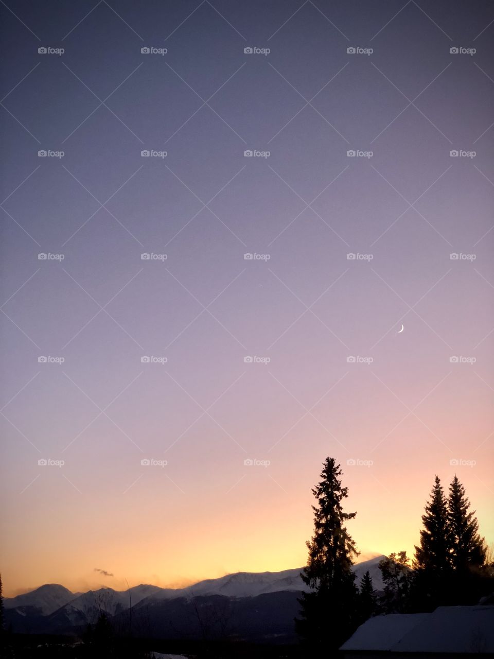 A sliver of a moon showing at sunset in the mountains with snow on the peaks.