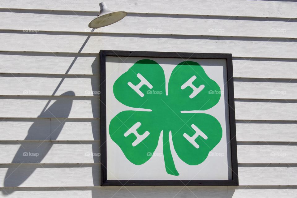 The 4-H logo on the side of a white wooden building with a vintage outdoor light casting a shadow