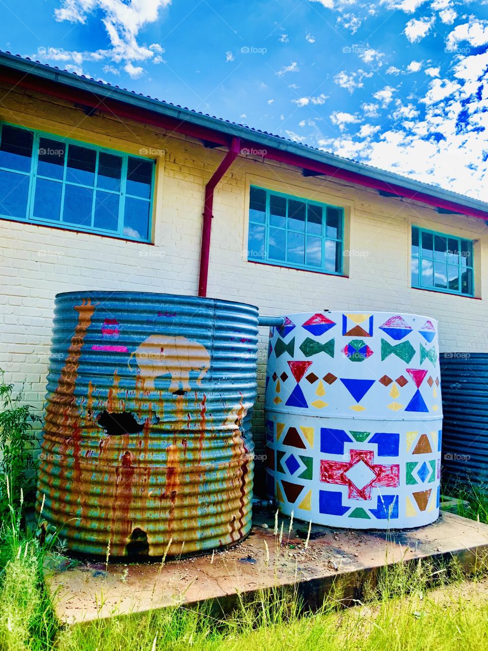 Colorfully painted plastic water tank, painted by children with geometric shapes and patterns with different bright colors; next to an old rusted metal tank behind a hostel block.