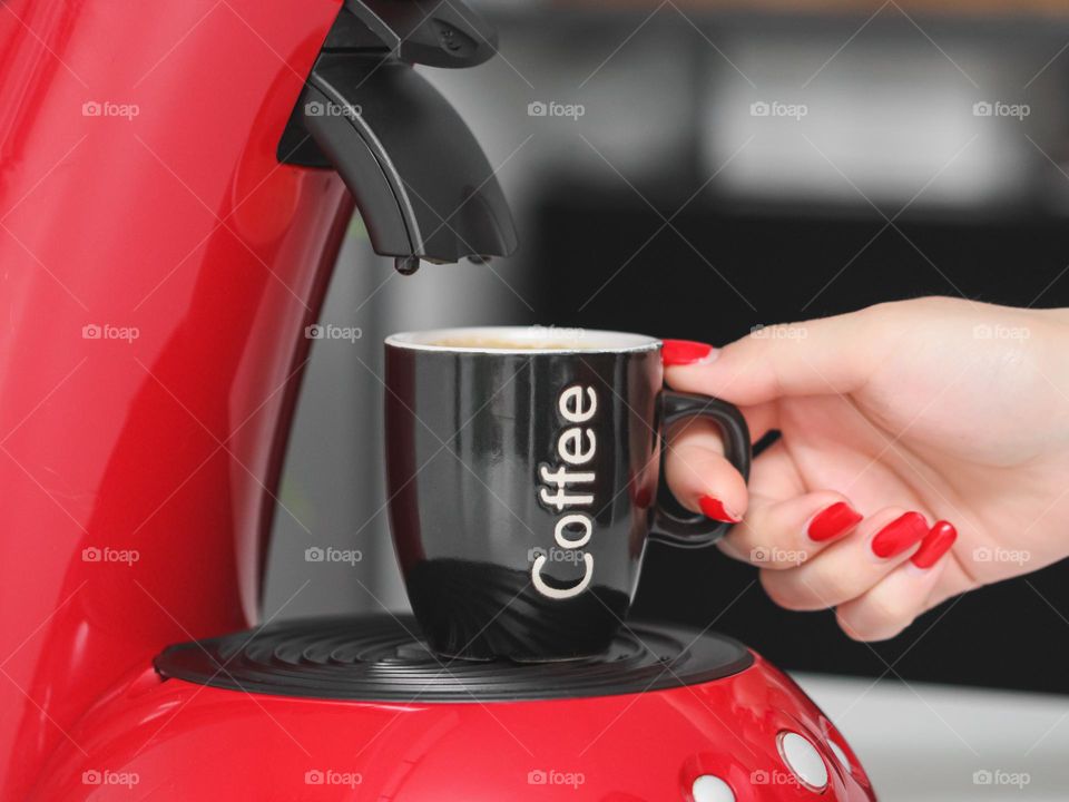 The hand of a young caucasian girl with a red manicure takes a black mug with the inscription: coffee from the coffee machine in the kitchen in the morning, side view close-up. The concept of morning drink, breakfast.