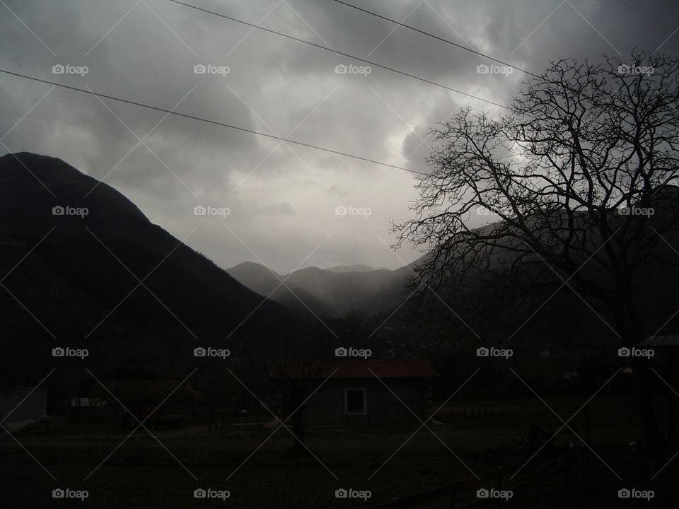 Cloudy afternoon in Morano Calabro, Italy. Evocative view of the sky on the mountains, a little country house in the middle and a beautiful bare tree on the right.