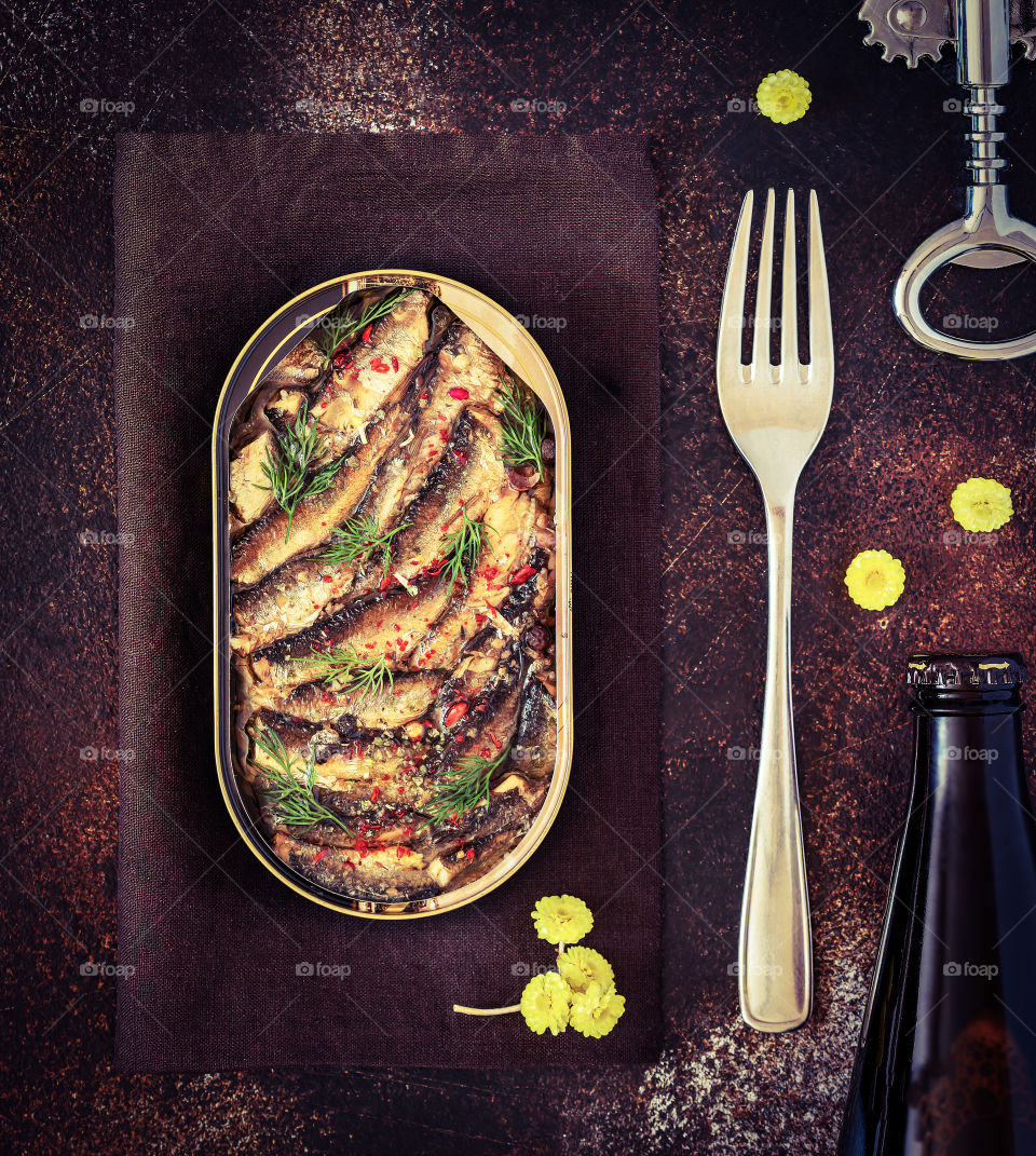 Open tin of Baltic sprats in oil with spices with a fork, a bottle of dark beer, a bottle opener and yellow flowers on an old brown background. Fish appetizer with beer. Food photo, top view