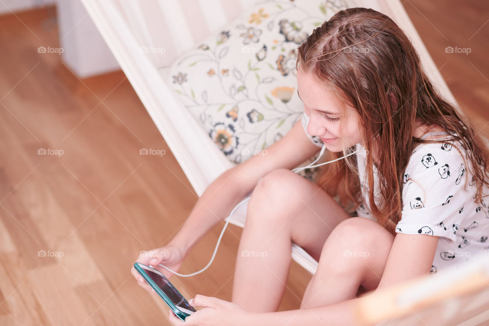 Teenage girl listening to music and chatting with friends on smartphone sitting in hammock at home
