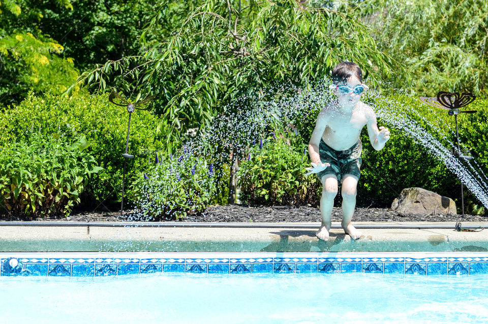 Young boy jumping through a sprinkler into a pool